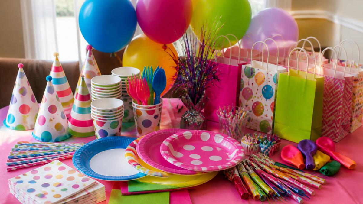 Colorful table display showing party supplies arranged together: themed paper plates, cups, napkins, utensils, balloons, decorations, party hats, and goodie bags in vibrant colors and themed designs